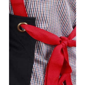 A black canvas bib apron with a red tie, featuring a grommet detail and a checkered fabric background.