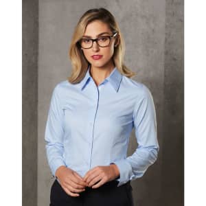 A woman wears a light blue long-sleeve button-up shirt, featuring a collar and a logo. The background is grey.