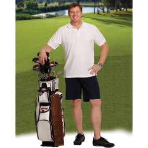 A man stands by a golf bag wearing navy chino shorts and a white polo shirt, with a lush green landscape in the background.