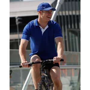 A man in a bright blue polo shirt and cap rides a bicycle, smiling. The polo features a logo.