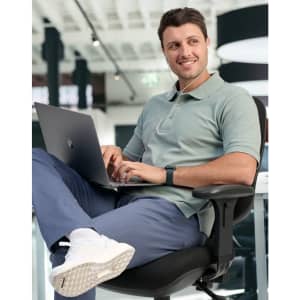 A men's cotton stretch polo in light green, featuring a logo, worn by a man seated at a desk with a laptop.