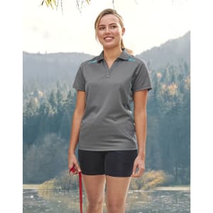 A woman stands outdoors wearing a grey polo shirt with blue contrast accents, alongside black shorts and holding a leash.