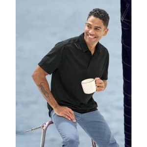 A man sits on a railing, wearing a black sustainable jacquard knit polo shirt and holding a white mug.
