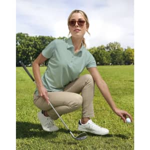 A light green polo shirt is worn by a woman crouching on grass, holding a golf ball, with sunglasses and casual trousers.