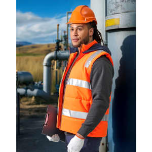 A man wearing an orange hi-vis hooded puffer vest stands beside a metallic structure, holding a laptop.