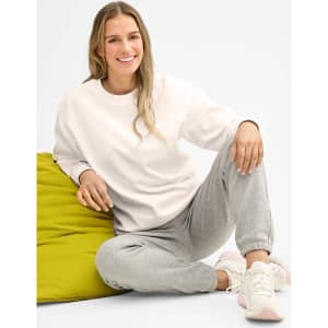 A woman sitting on a green bean bag, wearing grey sweatpants and a white sweatshirt, smiling at the camera.