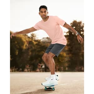 A man in a light pink premium cotton tee skates on a skateboard, wearing grey shorts and white sneakers.