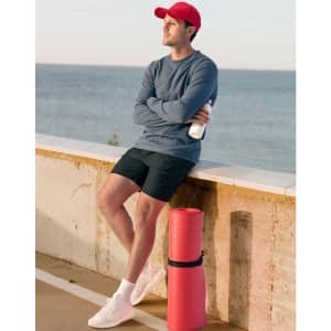 A man in a grey long-sleeve tee and red cap stands by a beach, holding a drink bottle next to a red yoga mat.