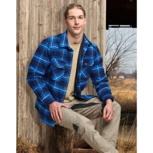 A young man wearing a blue and black quilted flannel shirt poses outdoors, seated against a wooden structure.