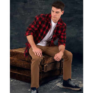 A man sits on a wooden crate wearing a red and black plaid flannel shirt over a white t-shirt with beige pants.