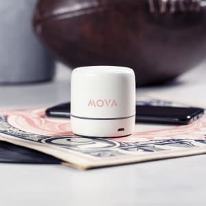 A compact white speaker sits on a table alongside a smartphone and some currency notes, with a brown football in the background.