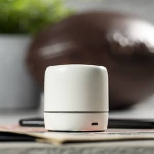 A white cylindrical speaker with a grey band, positioned on a table alongside a few items and a blurred object in the background.