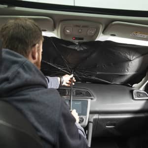 A black sunshade is being set up in a car's front window by a person in a grey hoodie.