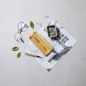 A bamboo pen box containing a pen, surrounded by paper clips, glasses, leaves, and a small container.