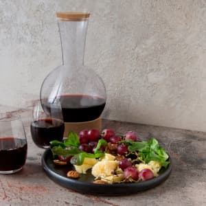 A wine decanter with a wooden stopper, filled with red wine, accompanied by two glasses and a plate of grapes and salad.