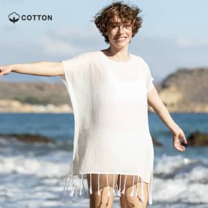 A light white kaftan with fringe detailing, modelled by a smiling person against a beach backdrop.