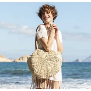 A large, round, natural-coloured tote bag with a fringed design, held by a smiling woman at the beach.