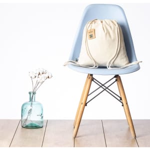 A cream drawstring bag with a logo sits on a light blue chair beside a glass bottle with cotton stems.