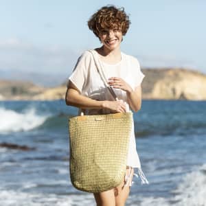 A woman smiling while holding a woven, gold tote bag by the beach, with waves in the background.