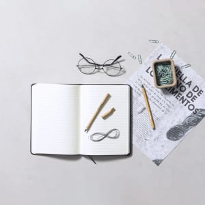 A flat lay featuring a notebook, two grey lead pencils, a pair of glasses, and a collection of paper clips on a light surface.