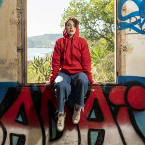 A red hooded sweatshirt is worn by a person sitting in a window, surrounded by greenery and graffiti.
