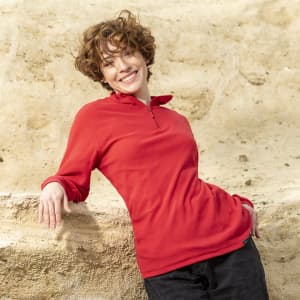 A red polar fleece jacket with a half zip and long sleeves, modelled by a smiling person against a sandy background.