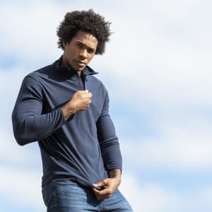 A man in a navy polar fleece jacket adjusts the collar against a cloudy sky backdrop, showcasing a stylish design.
