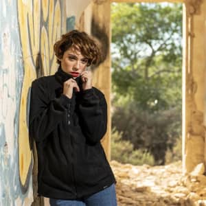 A black polar fleece jacket is worn by a model in an outdoor setting, against a graffiti-adorned wall.