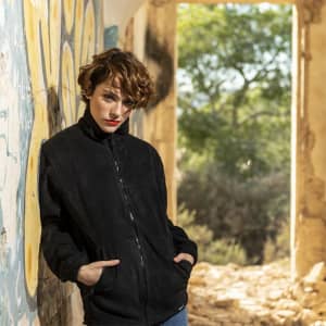A black polar fleece jacket worn by a person leaning against a graffiti-adorned wall, with greenery in the background.