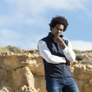 A man wearing a navy blue vest with a white long-sleeve shirt stands outdoors against a rocky backdrop.