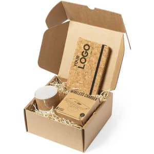 A brown gift box containing a cork notebook, wireless charger, and a drink bottle, set on shredded paper.