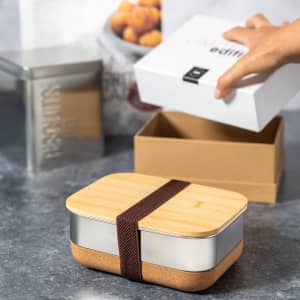 A bamboo and stainless steel lunch box with a brown elastic strap, placed on a textured surface beside boxes.