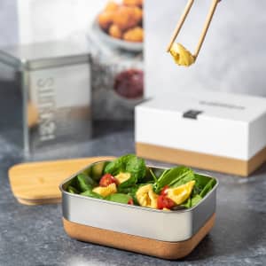 A lunch box with a silver lid containing spinach, cherry tomatoes, and pasta, with a wooden base and chopsticks.