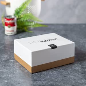 A white and brown lunch box with a logo, resting on a grey surface beside a can and some greenery.