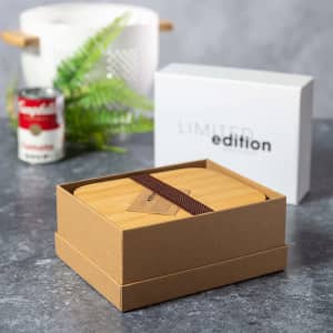 A bamboo lunch box in a kraft paper box with a brown strap, surrounded by greenery and a white container.