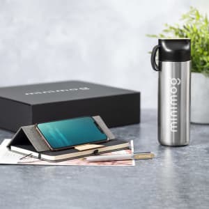 A stainless steel drink bottle, phone stand, notebook, and pen, arranged on a grey surface with a gift box.