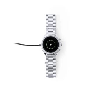 A silver smart watch with a circular face, charging with a cable attached, featuring a logo on the dial.
