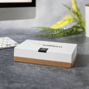 A white and brown box with a logo, placed on a grey surface surrounded by greenery and a computer keyboard.