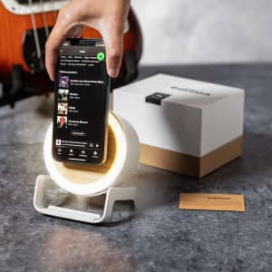 A multifunction lamp in white and wood, designed as a wireless charger, with a smartphone resting on it.