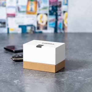 A rectangular box with a white top and brown base, featuring a logo, on a grey surface with phones in the background.