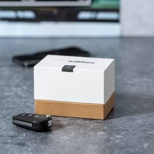 A white and brown box with a logo, placed on a grey surface beside a black car key and a smartphone.