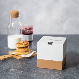 A square white box with a brown base, placed on a grey surface, next to a glass bottle and a plate of donuts.