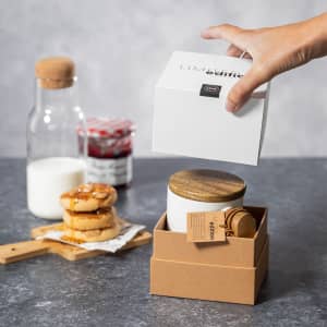 A coffee mug with a wooden lid in a gift box, alongside a glass bottle, jar of jam, cookies, and milk.