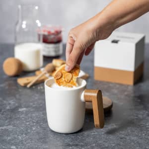 A white coffee mug with a wooden handle, accompanied by a jar, milk, and a snack being dipped into the mug.