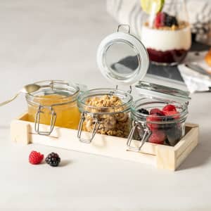 Three glass jars with metal clips, containing honey, granola, and mixed berries, placed on a wooden tray.