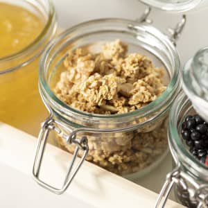 A glass jar filled with granola, featuring a metal clamp lid, beside other jars of preserves and fruit.