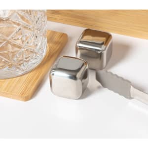 A drink bottle and two stainless steel ice cubes sit beside a beverage tongs on a light wooden surface.