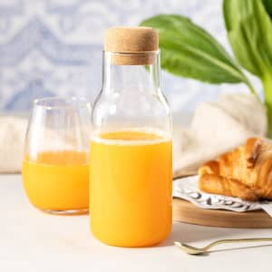A drink bottle made of clear glass with a cork lid, filled with orange juice, alongside a croissant and a glass.