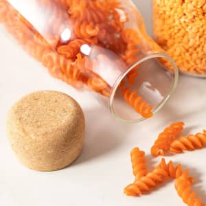 A drink bottle with a cork lid, featuring orange spiral pasta inside and surrounding it on a light background.