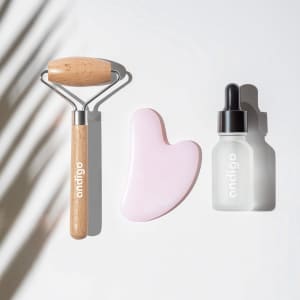 A pink face massager, a wooden roller, and a dropper bottle on a light surface. The items feature a logo.